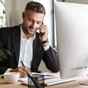 Image of handsome businessman 30s wearing suit talking on cell phone while working on computer in office